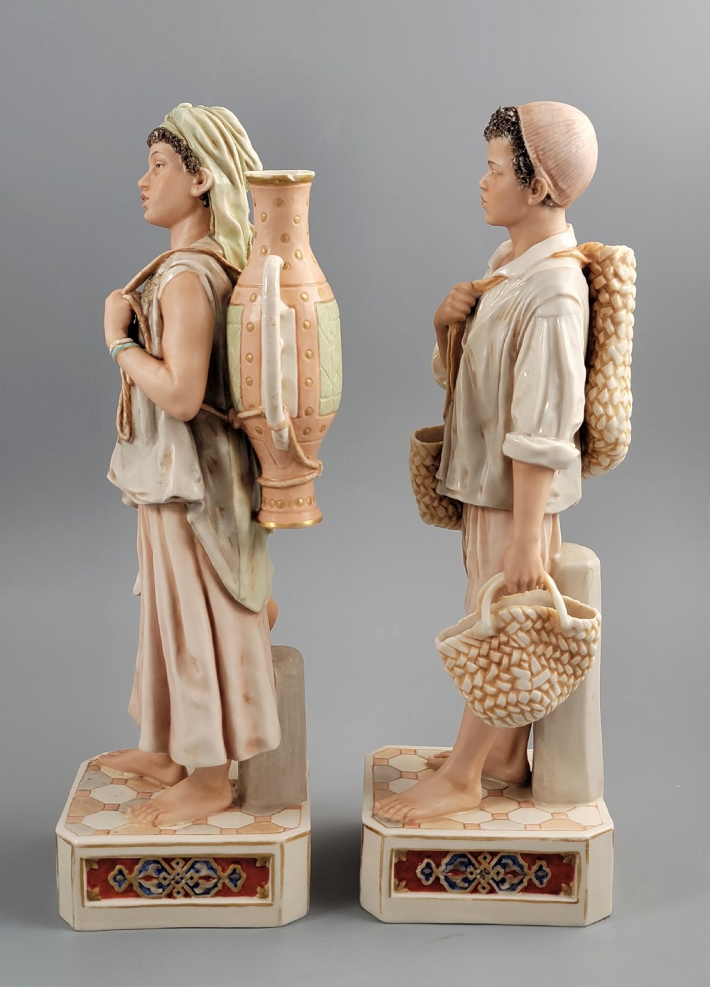 A pair of late 19th-century Royal Dux porcelain figurines depicting a male and female water bearer in draped attire standing on patterned pedestal bases. View II