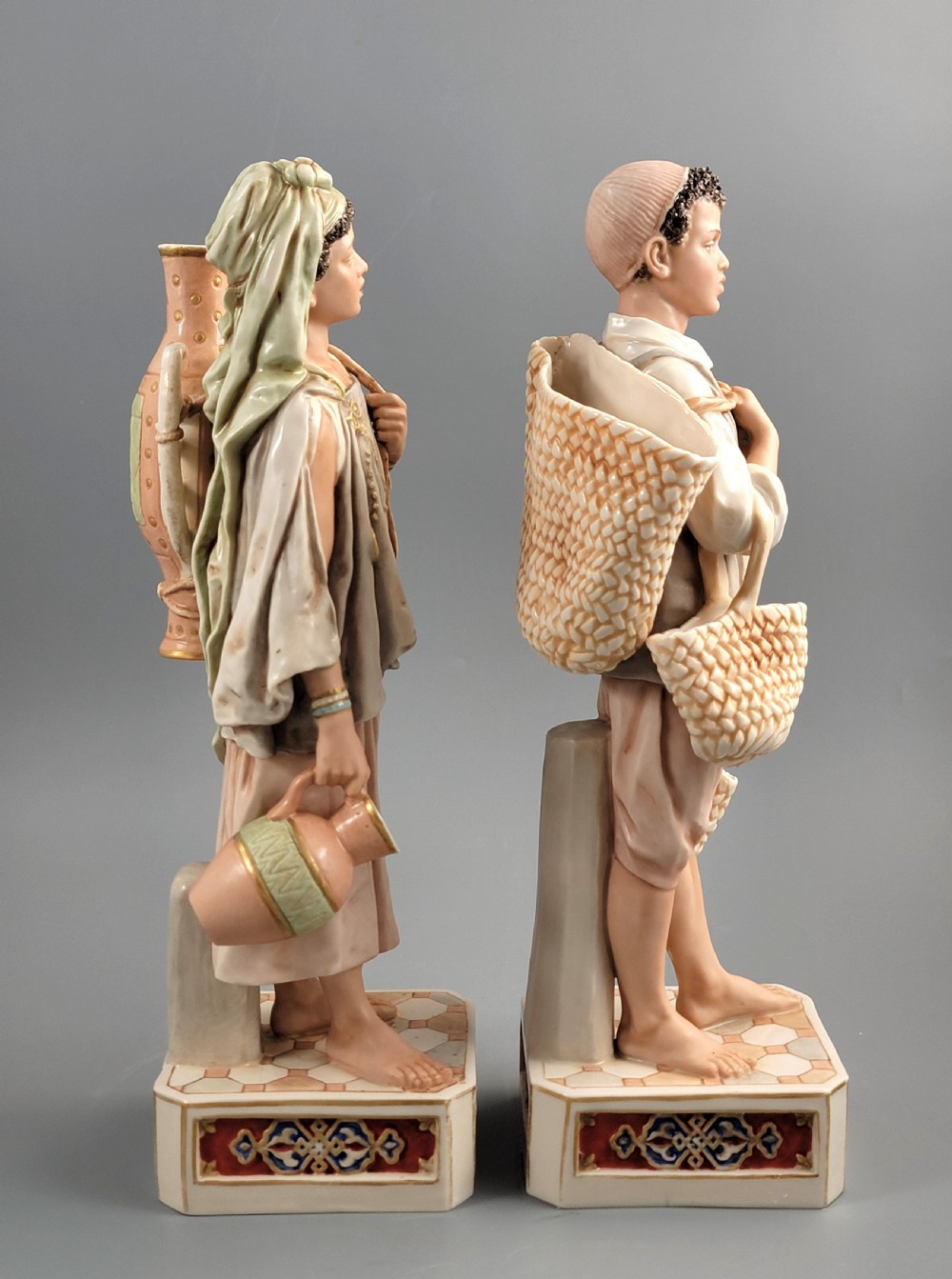 A pair of late 19th-century Royal Dux porcelain figurines depicting a male and female water bearer in draped attire standing on patterned pedestal bases. View IV