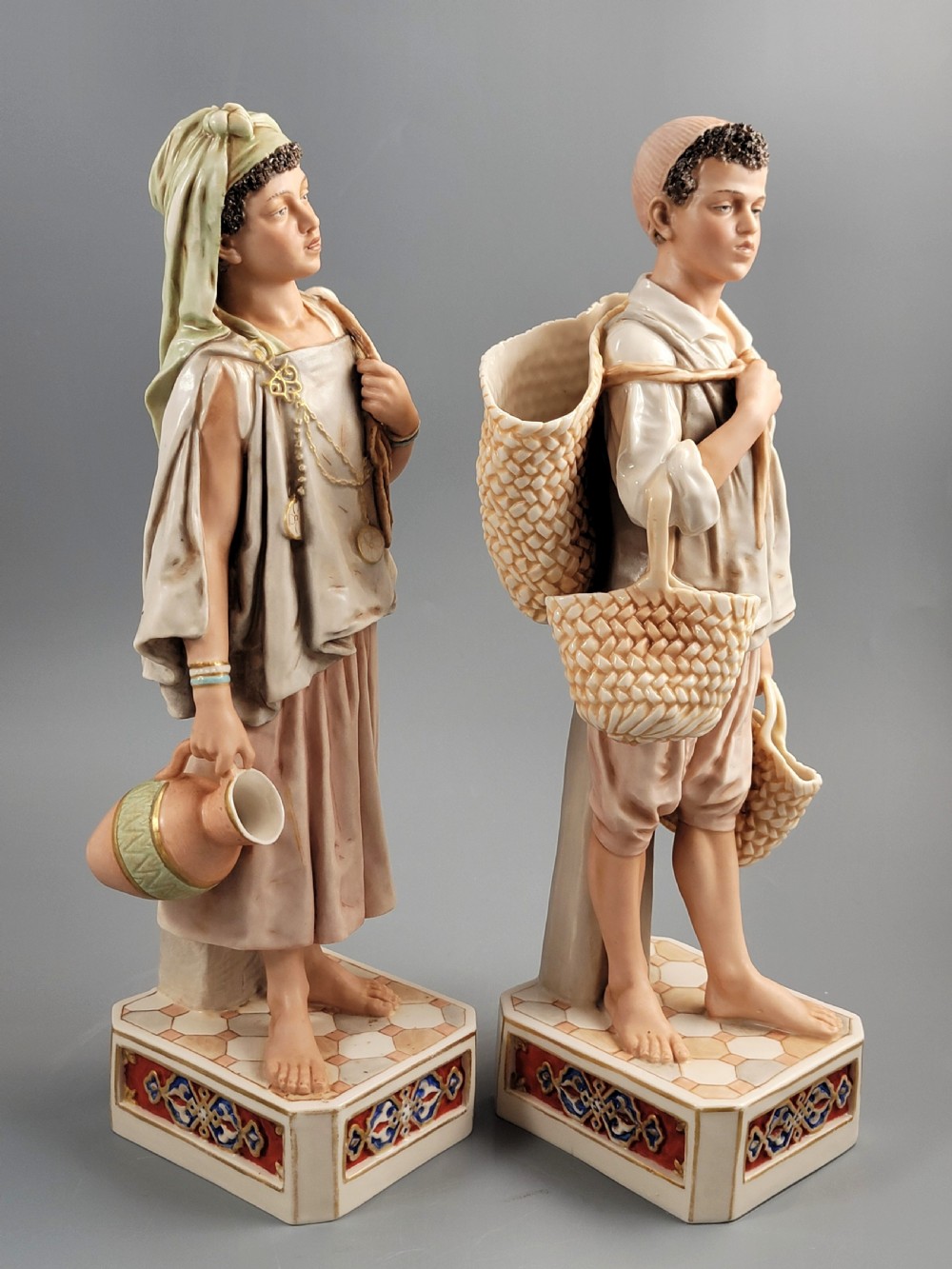 A pair of late 19th-century Royal Dux porcelain figurines depicting a male and female water bearer in draped attire standing on patterned pedestal bases. View V