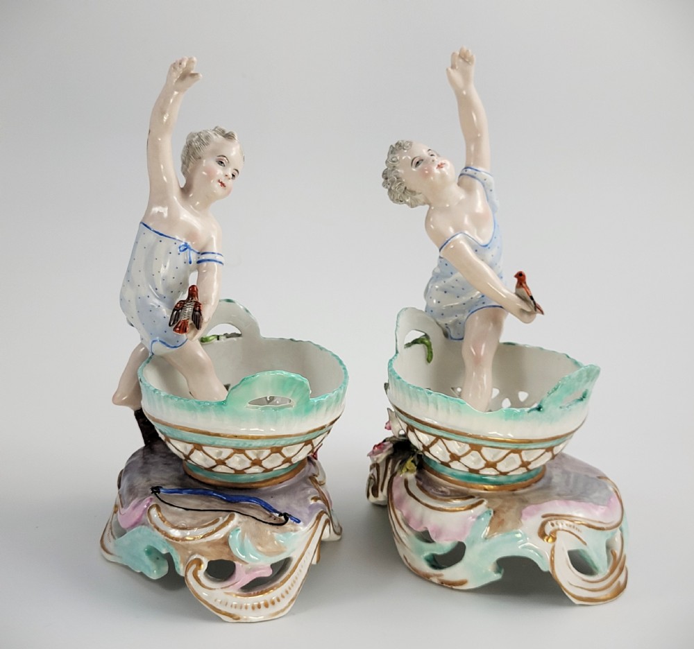 A pair of 19th-century German porcelain sweetmeat figures featuring sculpted children with birds beside reticulated baskets on ornate scrollwork bases.
