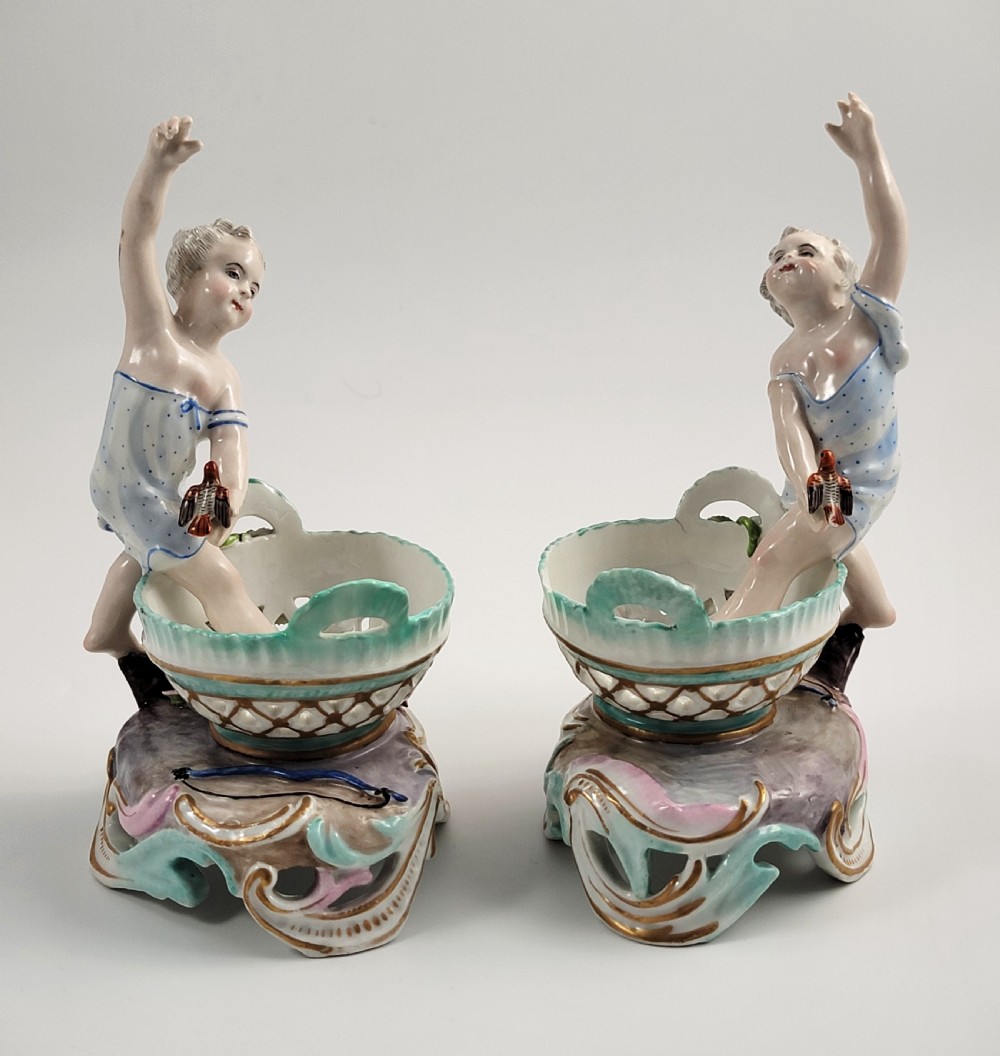 A pair of 19th-century German porcelain sweetmeat figures featuring sculpted children with birds beside reticulated baskets on ornate scrollwork bases. View II