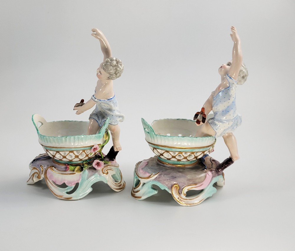 A pair of 19th-century German porcelain sweetmeat figures featuring sculpted children with birds beside reticulated baskets on ornate scrollwork bases. View IV