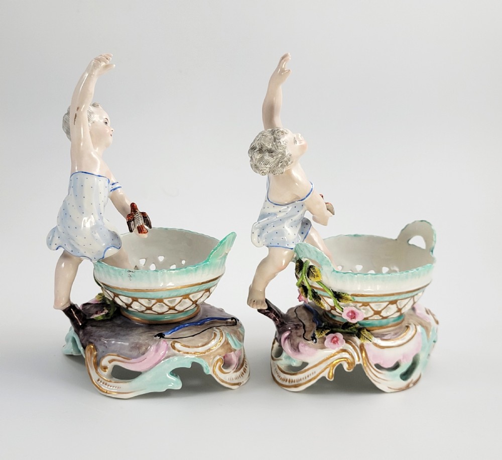 A pair of 19th-century German porcelain sweetmeat figures featuring sculpted children with birds beside reticulated baskets on ornate scrollwork bases. View V