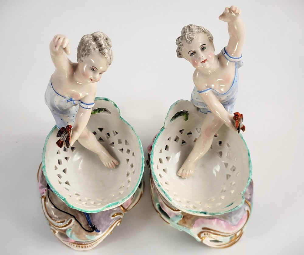A pair of 19th-century German porcelain sweetmeat figures featuring sculpted children with birds beside reticulated baskets on ornate scrollwork bases. View VI