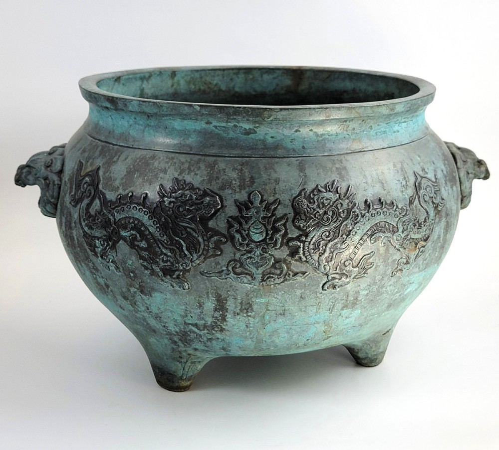 A tripod Chinese bronze censer decorated with raised dragon motifs, mask handles, and a verdigris surface finish.