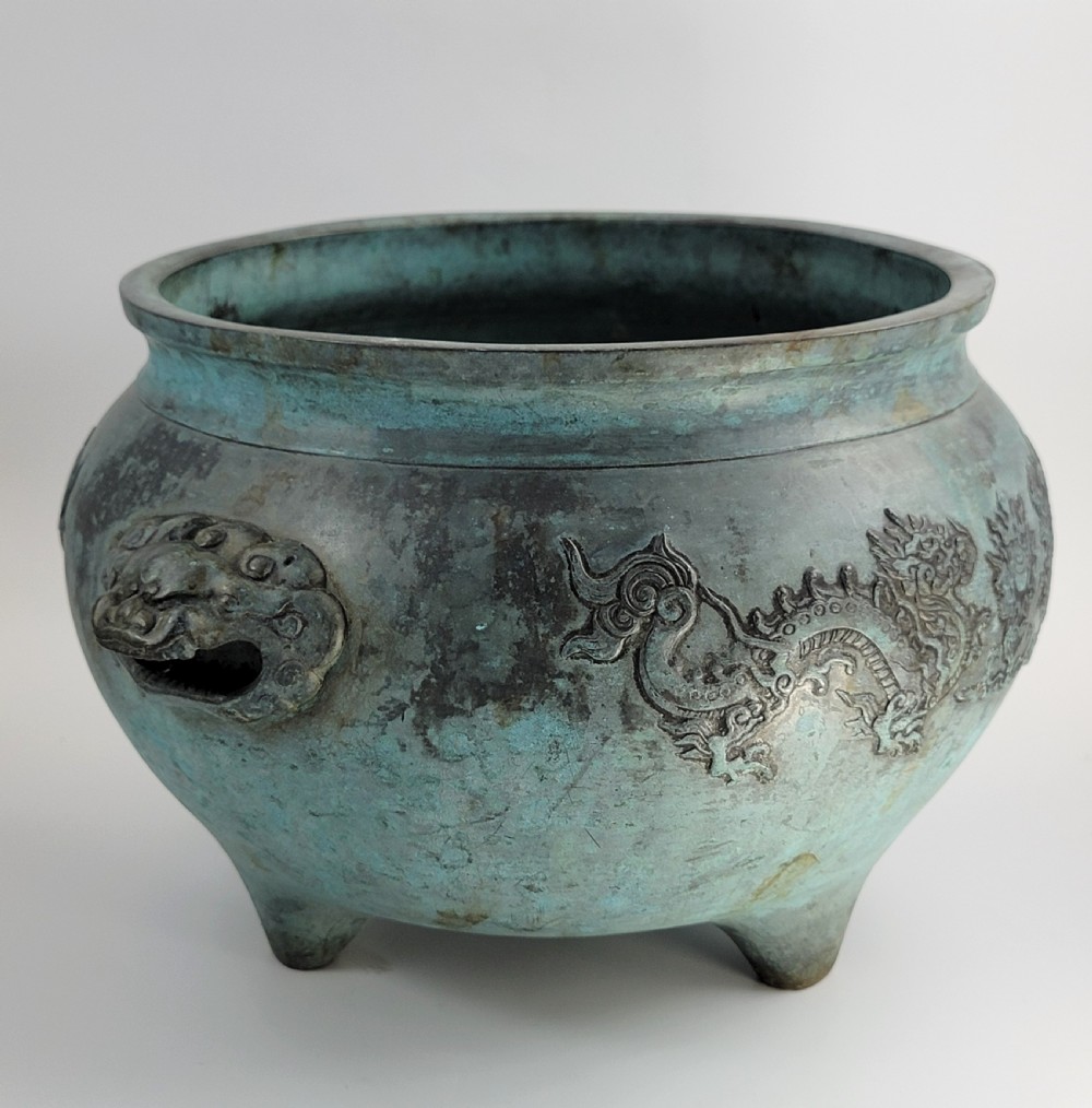 A tripod Chinese bronze censer decorated with raised dragon motifs, mask handles, and a verdigris surface finish. View V