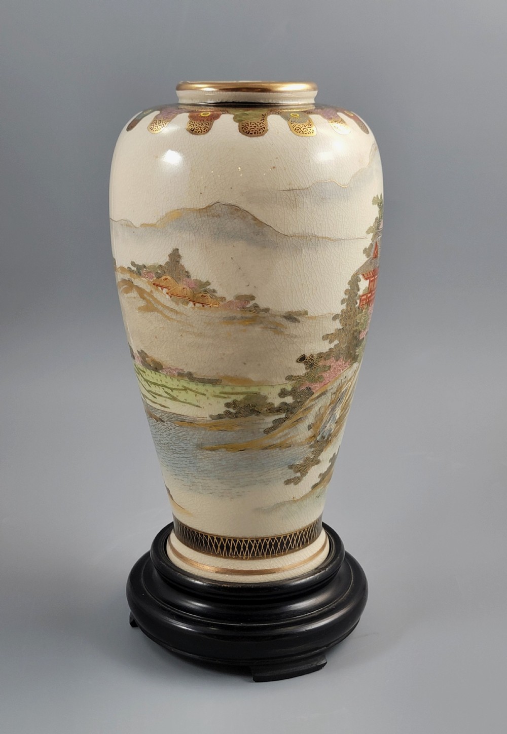An antique Japanese Satsuma earthenware vase decorated with hand-painted landscape scenes, figures, and pagodas over a fine crackle glaze, sitting on a turned dark wood stand. View V