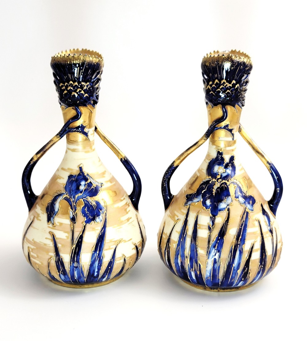 A pair of Art Nouveau porcelain vases featuring hand-painted cobalt blue iris flowers, gold-toned striped backgrounds, and gilded sweeping handles.