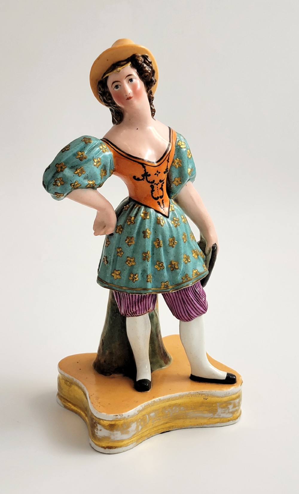 An early 19th-century French hard-paste porcelain figural scent bottle depicting a standing woman in a teal patterned dress, orange bodice, and yellow hat, mounted on a gilded shaped base.