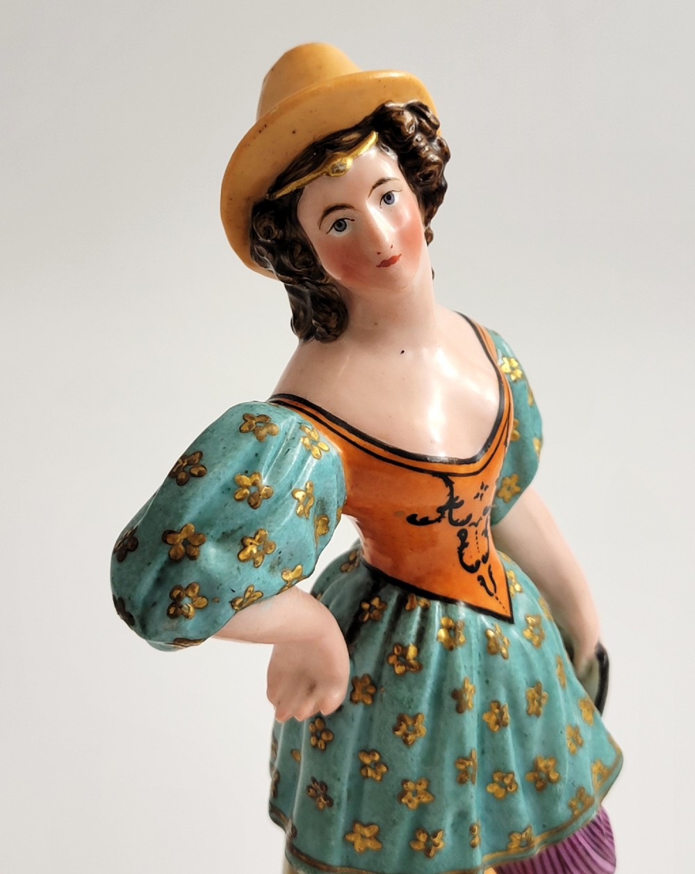 An early 19th-century French hard-paste porcelain figural scent bottle depicting a standing woman in a teal patterned dress, orange bodice, and yellow hat, mounted on a gilded shaped base. View VII