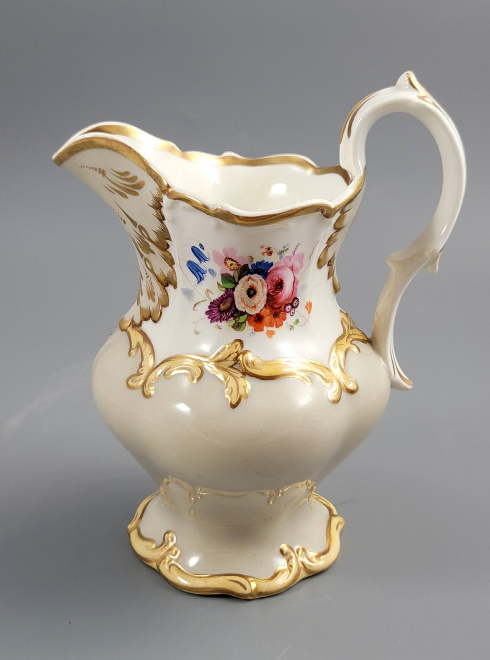 A white porcelain jug decorated with a hand-painted floral spray, gilded acanthus leaf scrollwork, a shaped spout, and a looped handle.