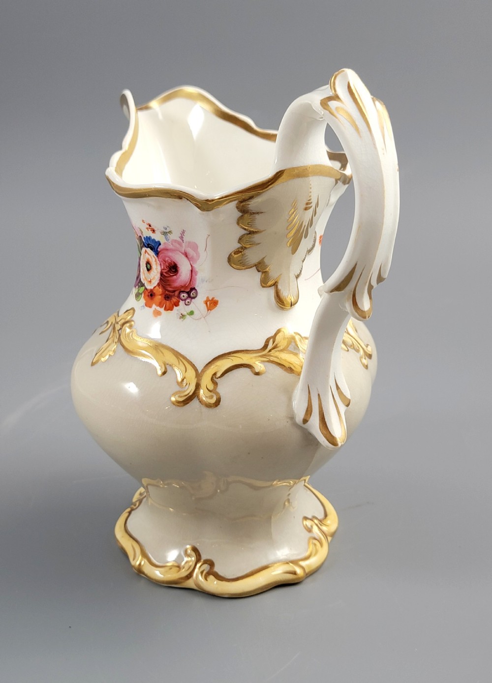 A white porcelain jug decorated with a hand-painted floral spray, gilded acanthus leaf scrollwork, a shaped spout, and a looped handle. View II