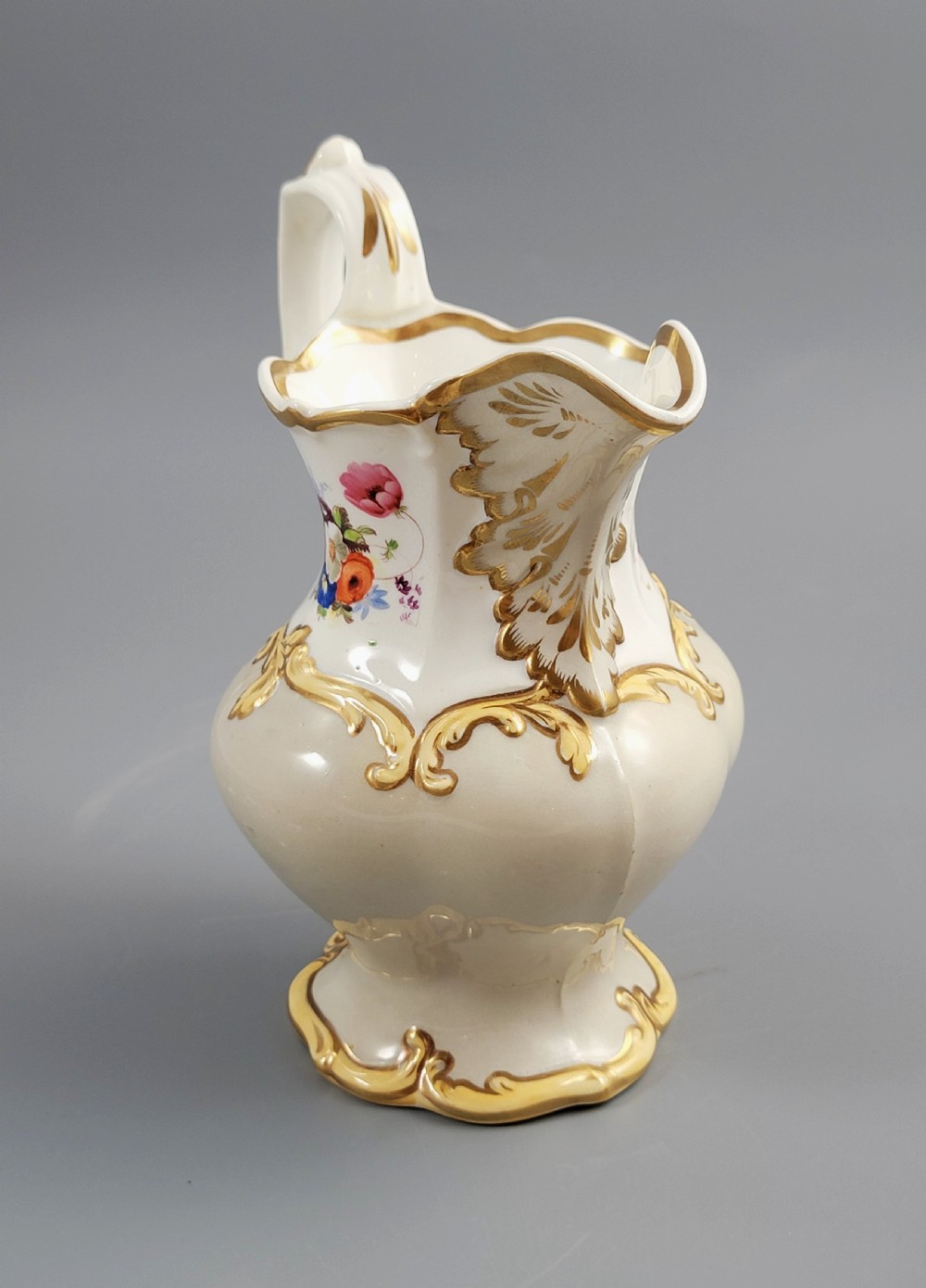 A white porcelain jug decorated with a hand-painted floral spray, gilded acanthus leaf scrollwork, a shaped spout, and a looped handle. View V