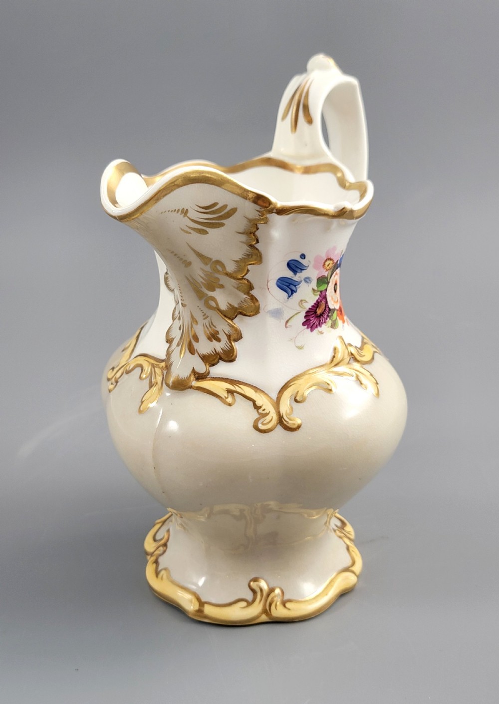 A white porcelain jug decorated with a hand-painted floral spray, gilded acanthus leaf scrollwork, a shaped spout, and a looped handle. View VI