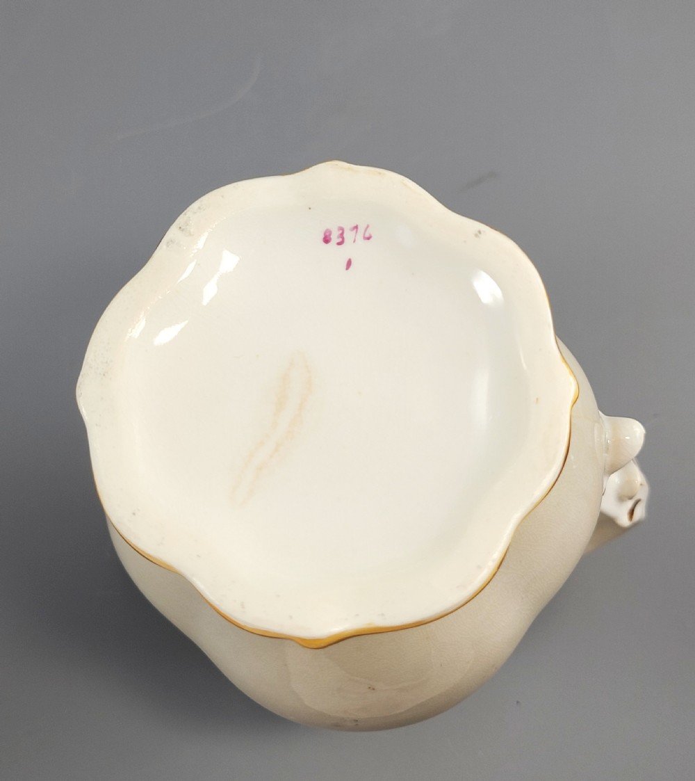 A white porcelain jug decorated with a hand-painted floral spray, gilded acanthus leaf scrollwork, a shaped spout, and a looped handle. View X