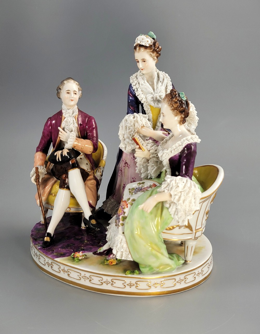 A German Volkstedt porcelain group depicting three eighteenth-century figures in elaborate attire seated on an oval gilt-decorated base. View II