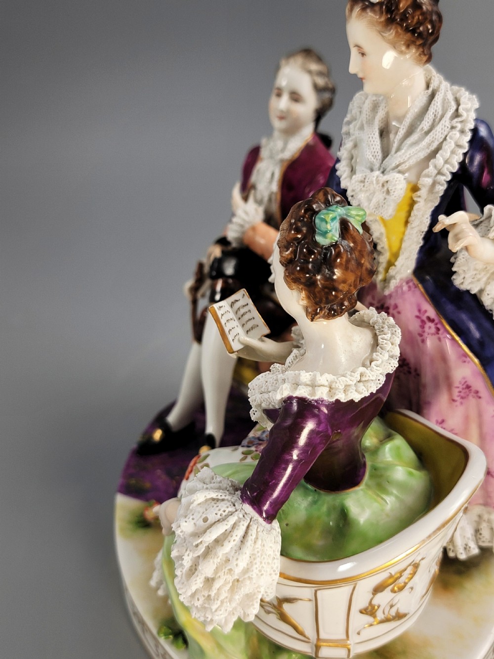 A German Volkstedt porcelain group depicting three eighteenth-century figures in elaborate attire seated on an oval gilt-decorated base. View X