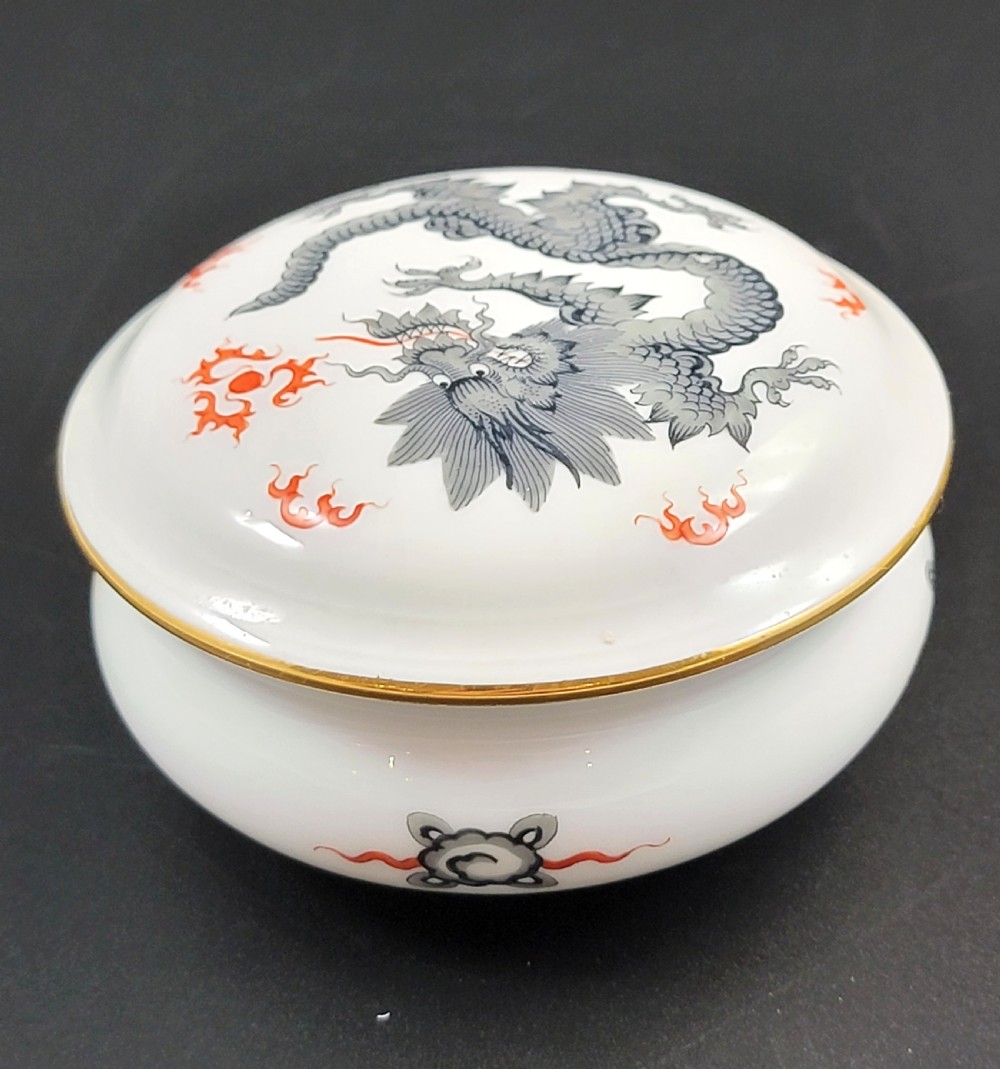 A circular white porcelain lidded trinket box adorned with a hand-painted grey dragon motif, red flame accents, and a gold gilt rim. View II
