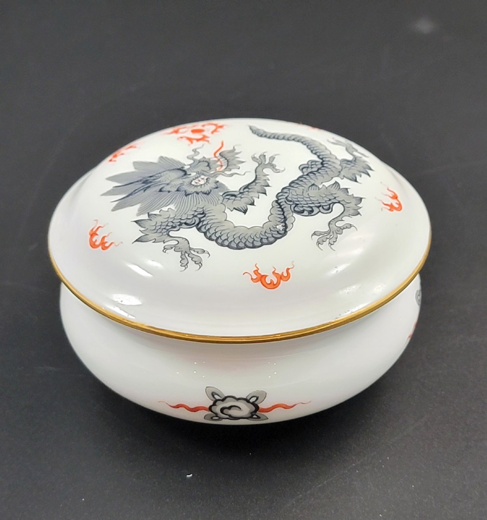 A circular white porcelain lidded trinket box adorned with a hand-painted grey dragon motif, red flame accents, and a gold gilt rim. View III