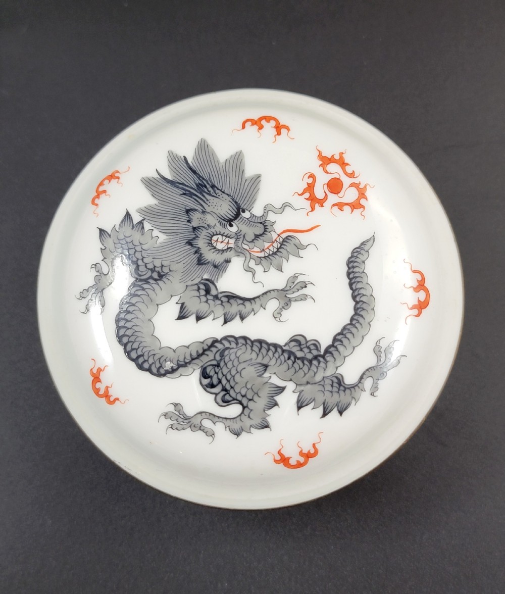 A circular white porcelain lidded trinket box adorned with a hand-painted grey dragon motif, red flame accents, and a gold gilt rim. View V