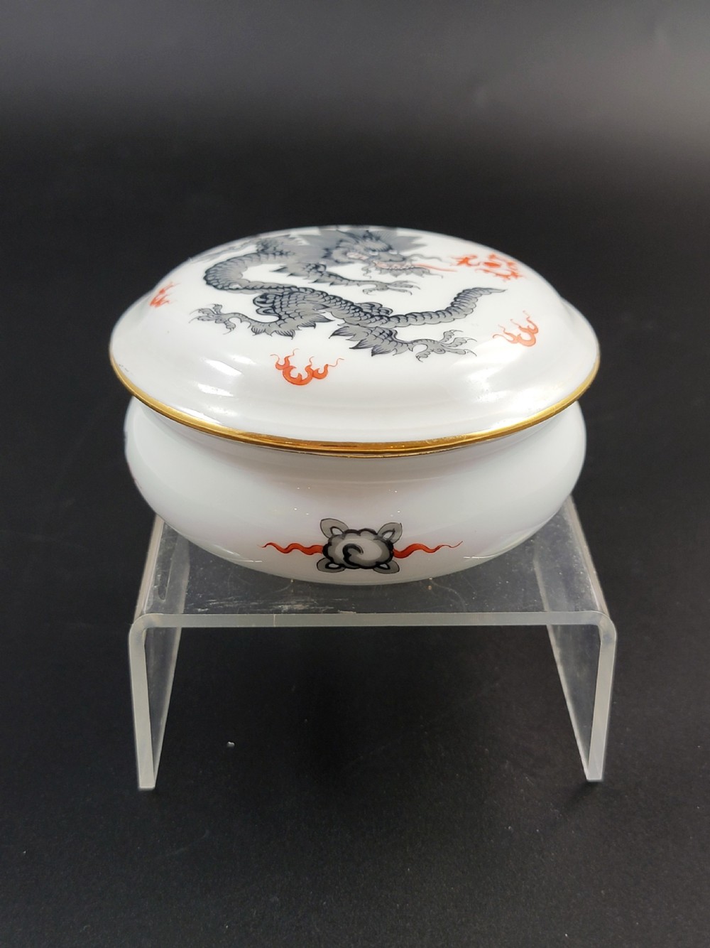 A circular white porcelain lidded trinket box adorned with a hand-painted grey dragon motif, red flame accents, and a gold gilt rim. View X