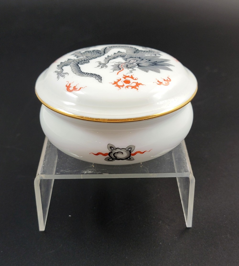 A circular white porcelain lidded trinket box adorned with a hand-painted grey dragon motif, red flame accents, and a gold gilt rim. View XI