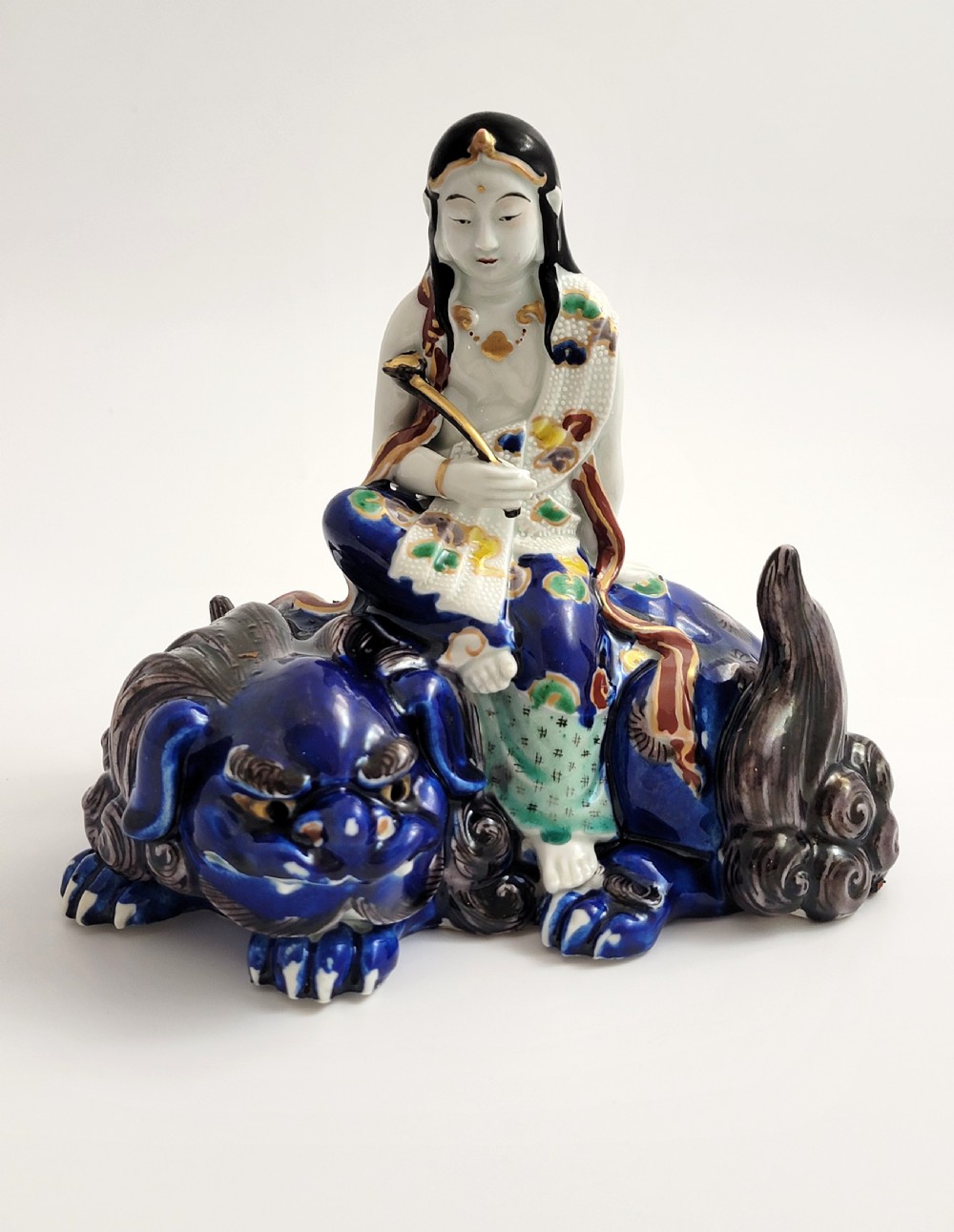 A Japanese Kutani porcelain figure depicting the deity Kannon seated atop a reclined blue shishi lion with detailed polychrome enamelled robes.