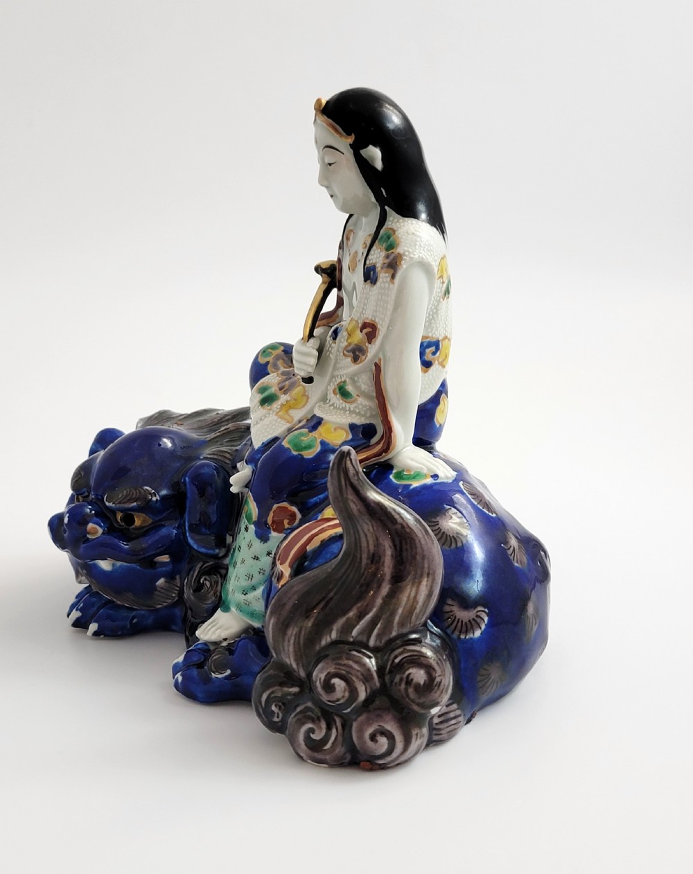 A Japanese Kutani porcelain figure depicting the deity Kannon seated atop a reclined blue shishi lion with detailed polychrome enamelled robes. View II