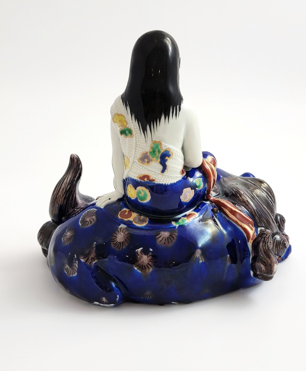 A Japanese Kutani porcelain figure depicting the deity Kannon seated atop a reclined blue shishi lion with detailed polychrome enamelled robes. View IV
