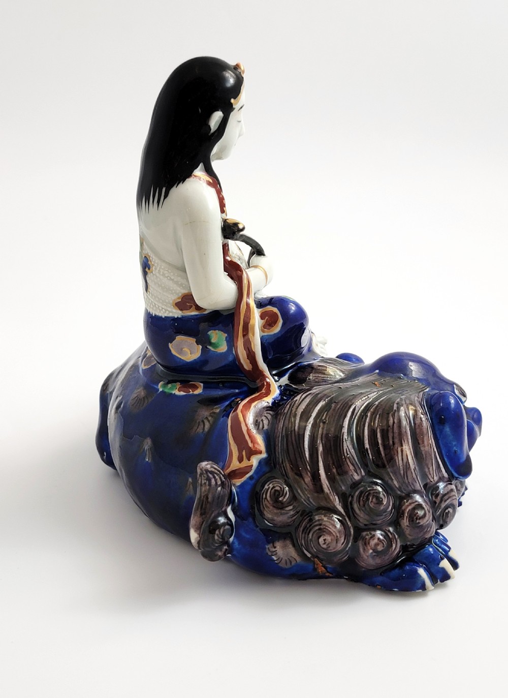 A Japanese Kutani porcelain figure depicting the deity Kannon seated atop a reclined blue shishi lion with detailed polychrome enamelled robes. View V