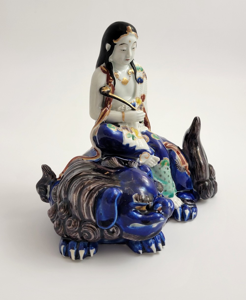 A Japanese Kutani porcelain figure depicting the deity Kannon seated atop a reclined blue shishi lion with detailed polychrome enamelled robes. View VI