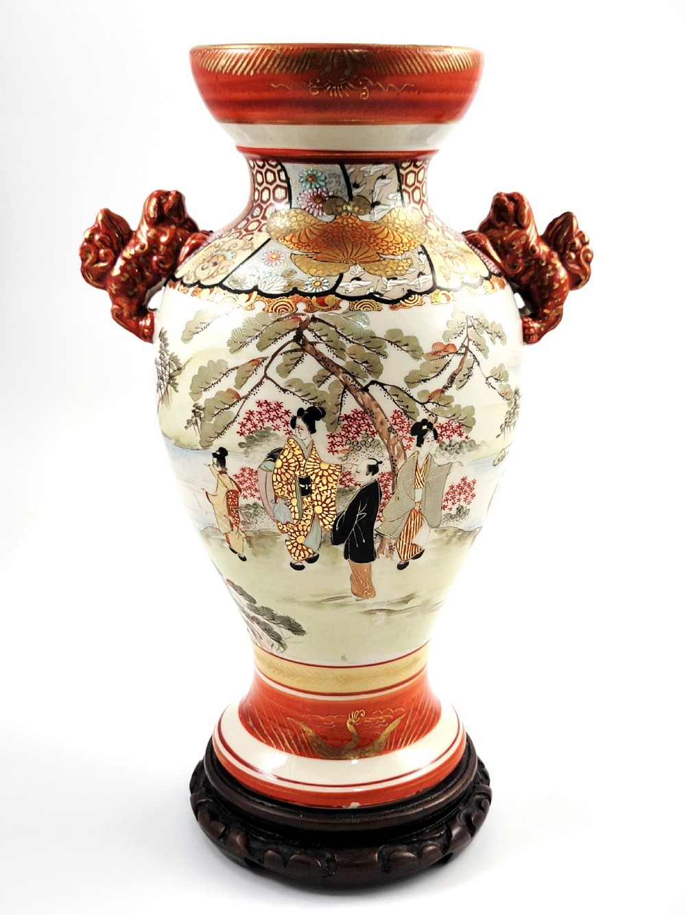 An antique Japanese Kutani porcelain vase featuring hand-painted figures, iron-red flared rim, gilded highlights, moulded lion handles, and a carved wooden display stand.