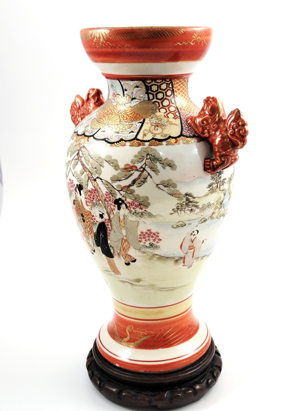 An antique Japanese Kutani porcelain vase featuring hand-painted figures, iron-red flared rim, gilded highlights, moulded lion handles, and a carved wooden display stand. View II