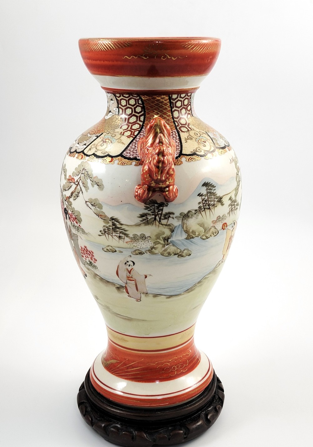 An antique Japanese Kutani porcelain vase featuring hand-painted figures, iron-red flared rim, gilded highlights, moulded lion handles, and a carved wooden display stand. View III