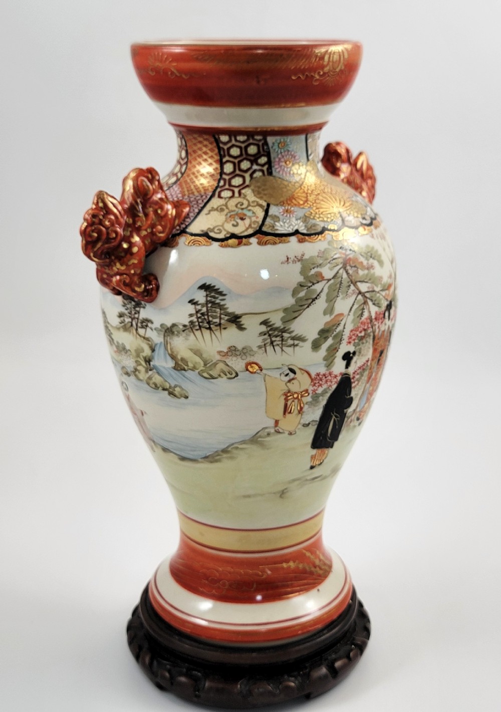 An antique Japanese Kutani porcelain vase featuring hand-painted figures, iron-red flared rim, gilded highlights, moulded lion handles, and a carved wooden display stand. View IV