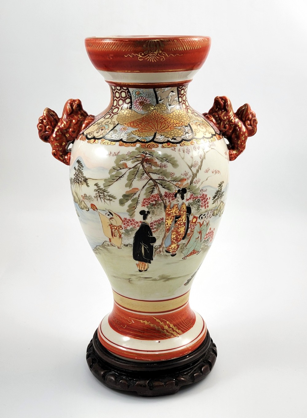 An antique Japanese Kutani porcelain vase featuring hand-painted figures, iron-red flared rim, gilded highlights, moulded lion handles, and a carved wooden display stand. View V