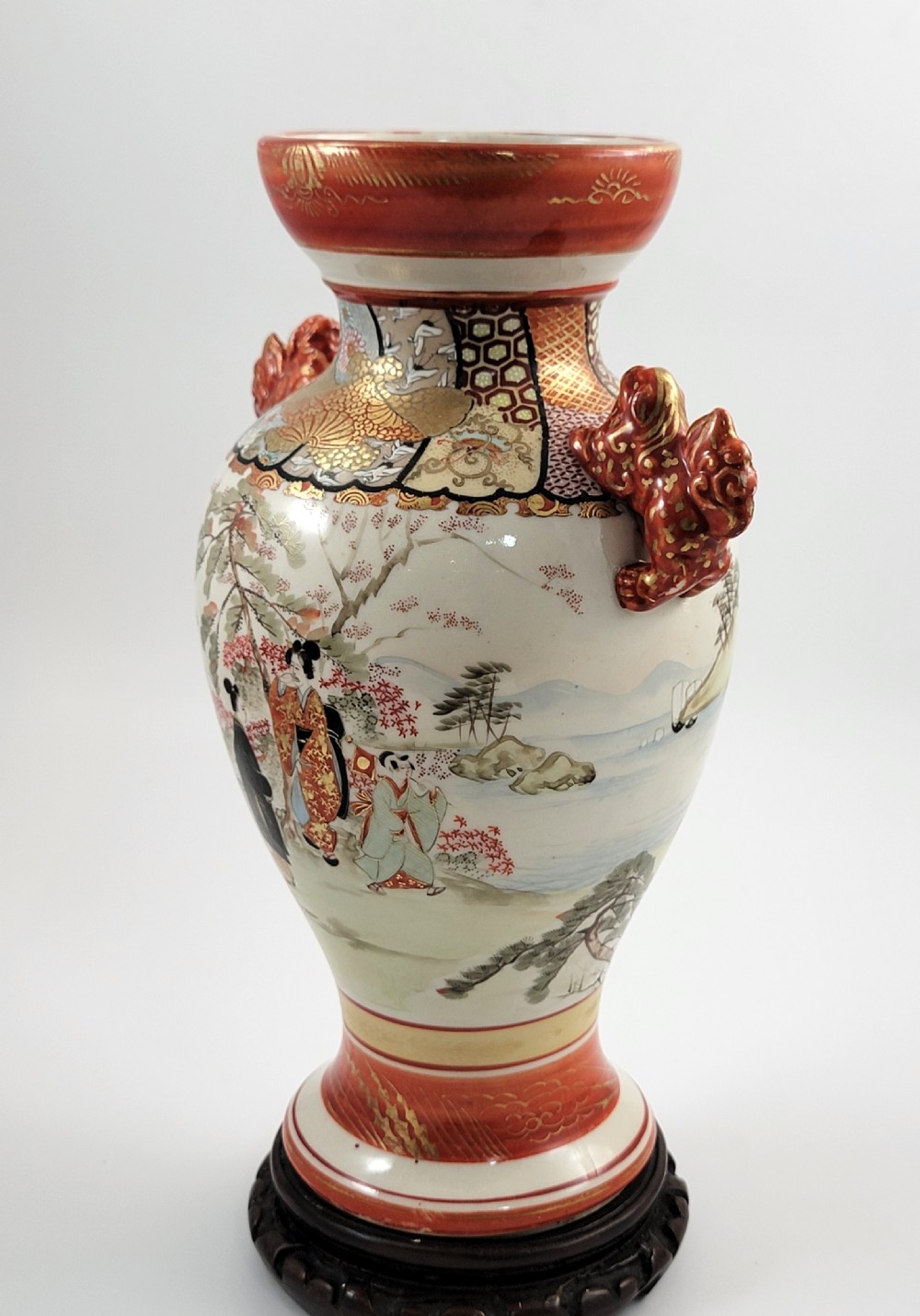 An antique Japanese Kutani porcelain vase featuring hand-painted figures, iron-red flared rim, gilded highlights, moulded lion handles, and a carved wooden display stand. View VI