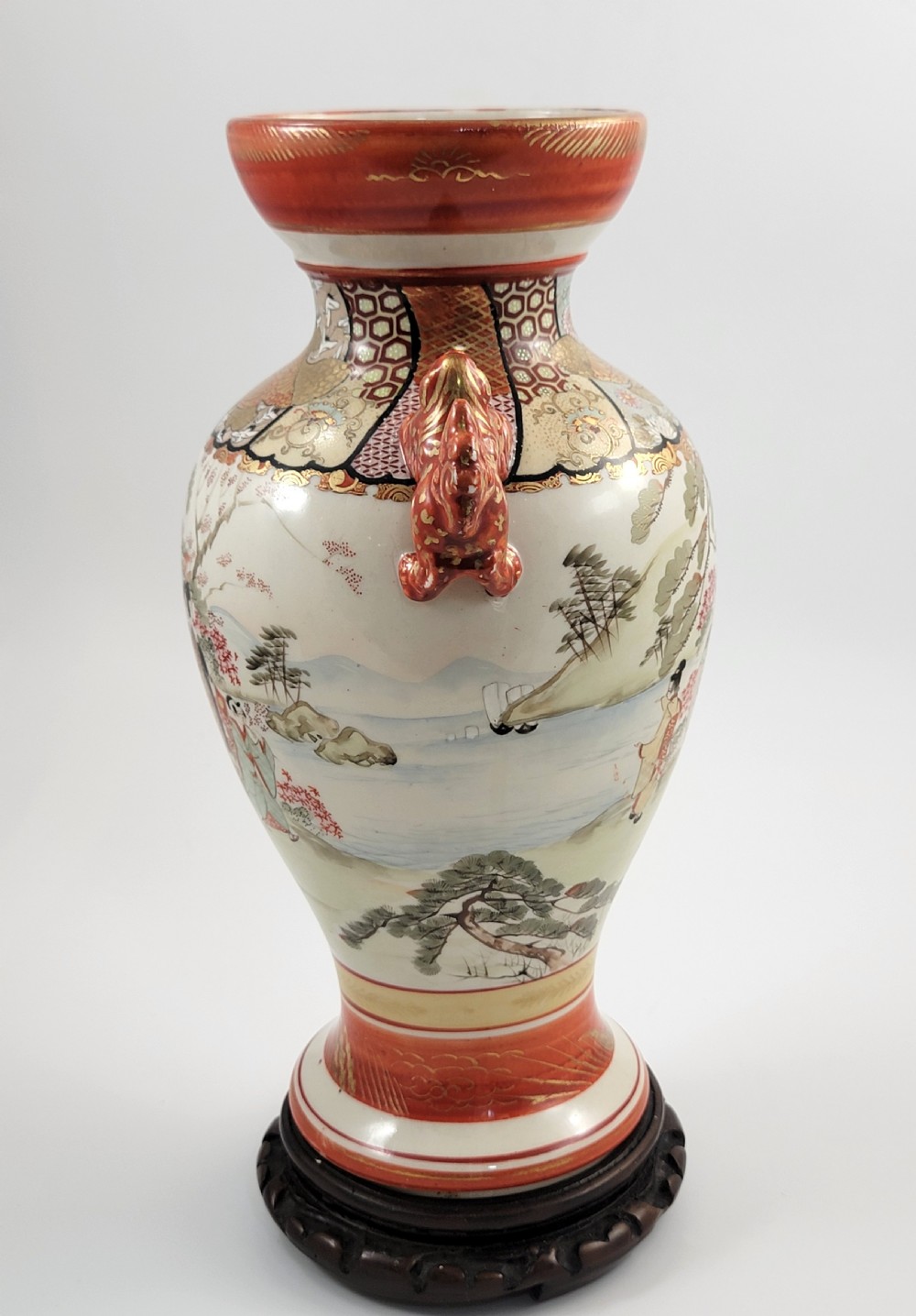 An antique Japanese Kutani porcelain vase featuring hand-painted figures, iron-red flared rim, gilded highlights, moulded lion handles, and a carved wooden display stand. View VII