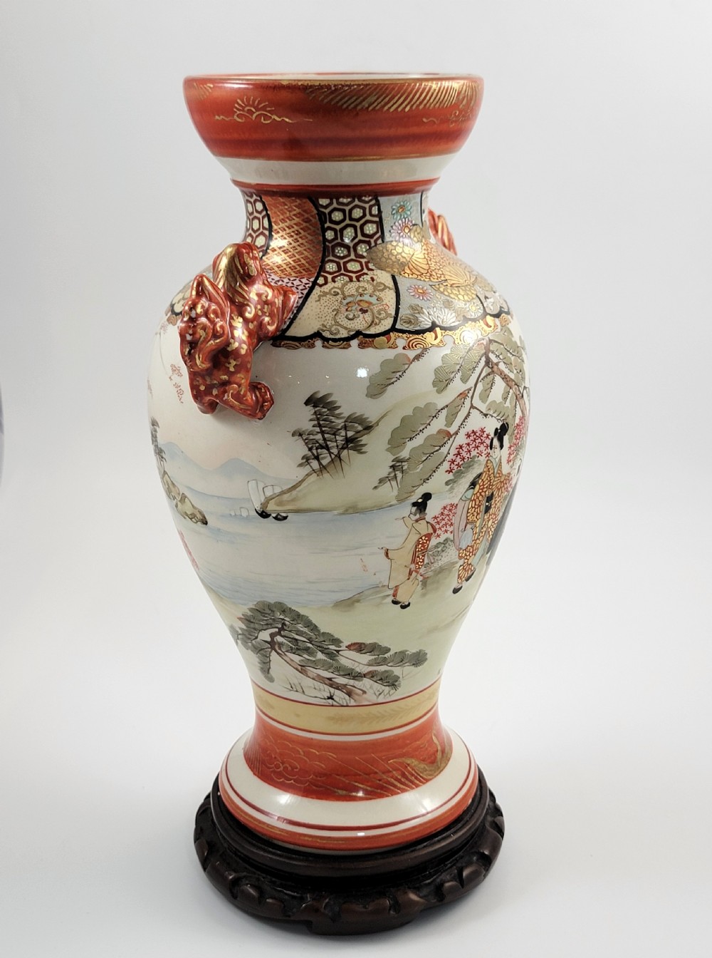 An antique Japanese Kutani porcelain vase featuring hand-painted figures, iron-red flared rim, gilded highlights, moulded lion handles, and a carved wooden display stand. View VIII
