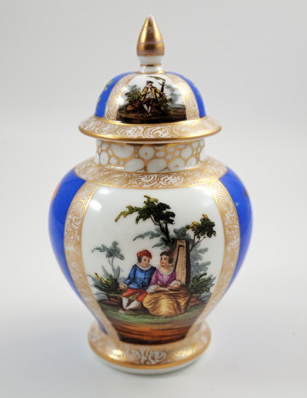 A lidded Dresden porcelain ginger jar featuring hand-painted courting scenes inside gilded cartouches against a cobalt blue ground, topped with a pointed gilt finial.