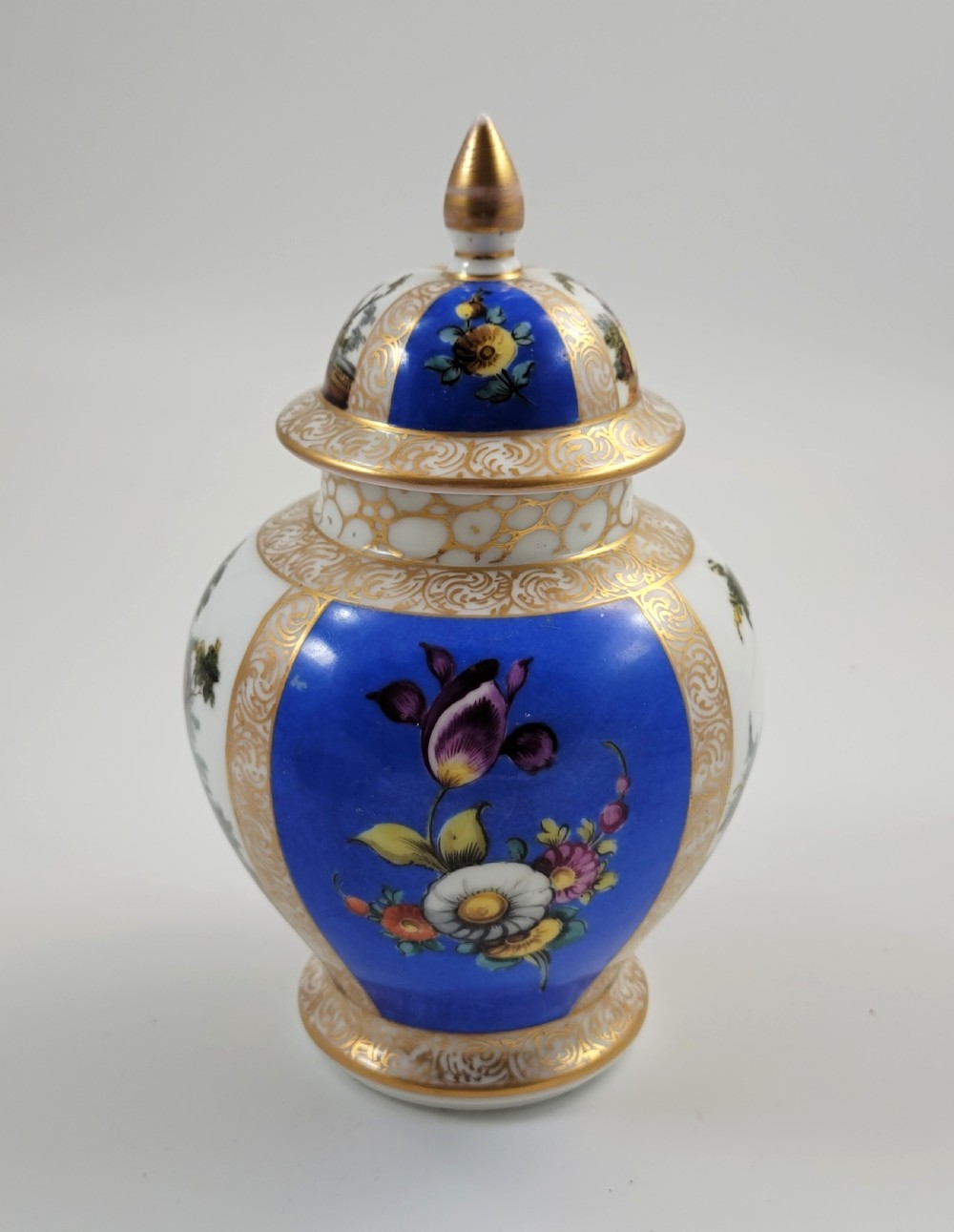 A lidded Dresden porcelain ginger jar featuring hand-painted courting scenes inside gilded cartouches against a cobalt blue ground, topped with a pointed gilt finial. View II