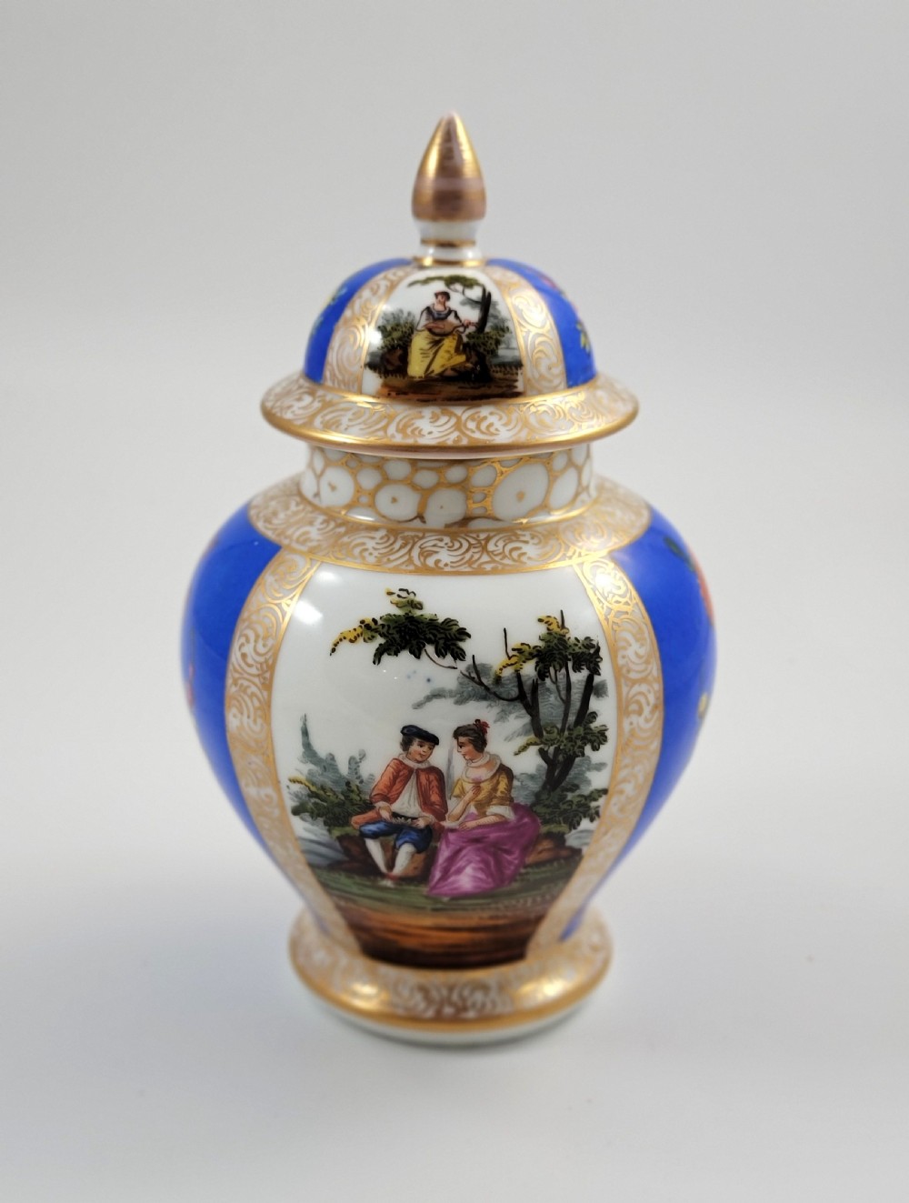 A lidded Dresden porcelain ginger jar featuring hand-painted courting scenes inside gilded cartouches against a cobalt blue ground, topped with a pointed gilt finial. View III