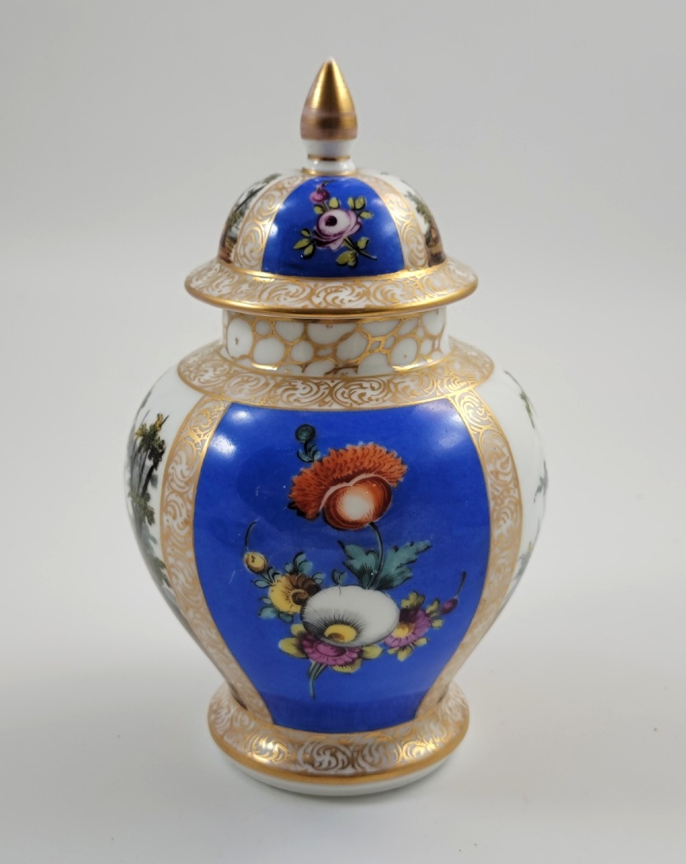 A lidded Dresden porcelain ginger jar featuring hand-painted courting scenes inside gilded cartouches against a cobalt blue ground, topped with a pointed gilt finial. View IV
