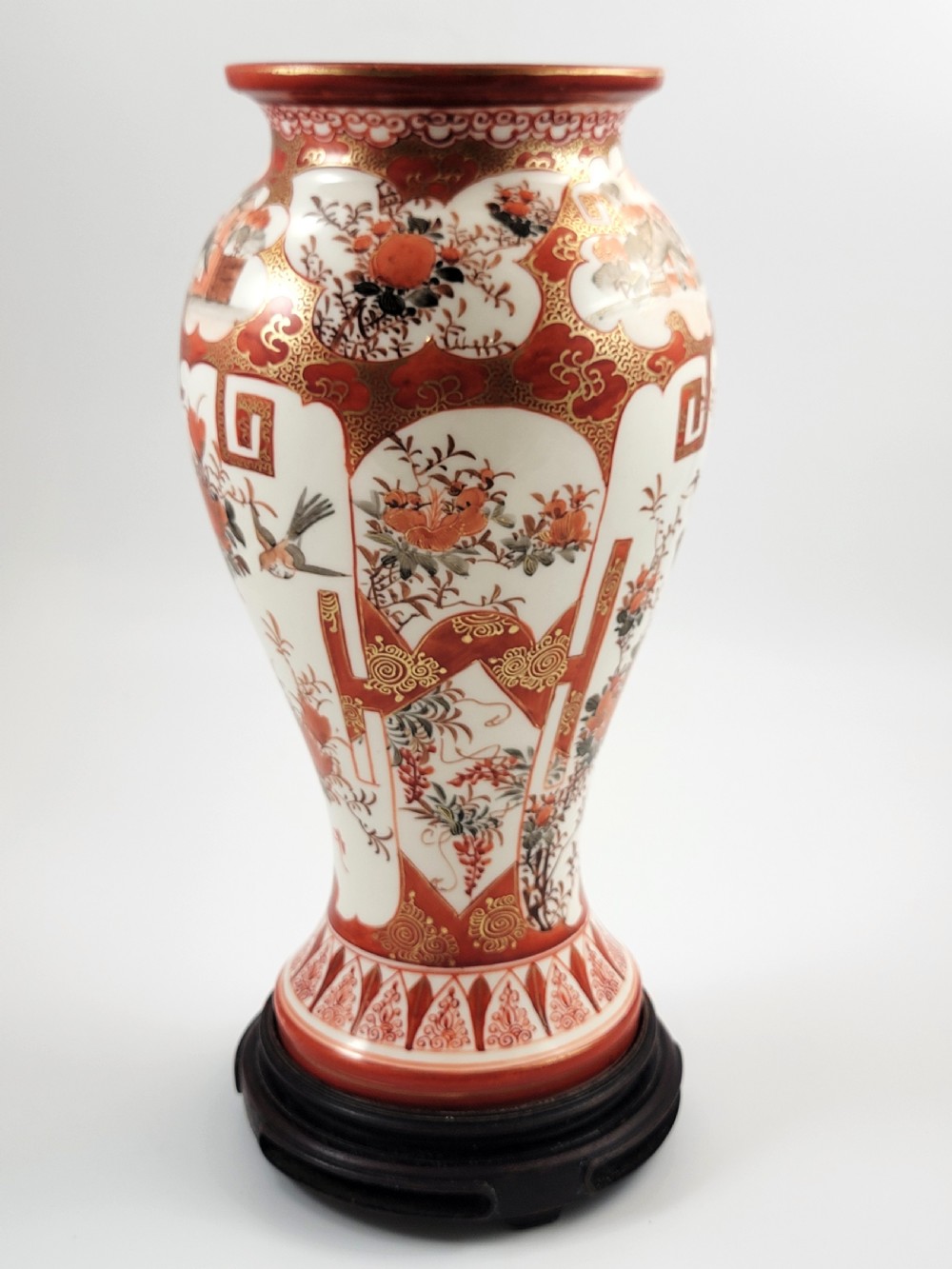 A baluster-shaped Japanese Kutani porcelain vase decorated with red and gold floral motifs on a carved dark wood base. View VI