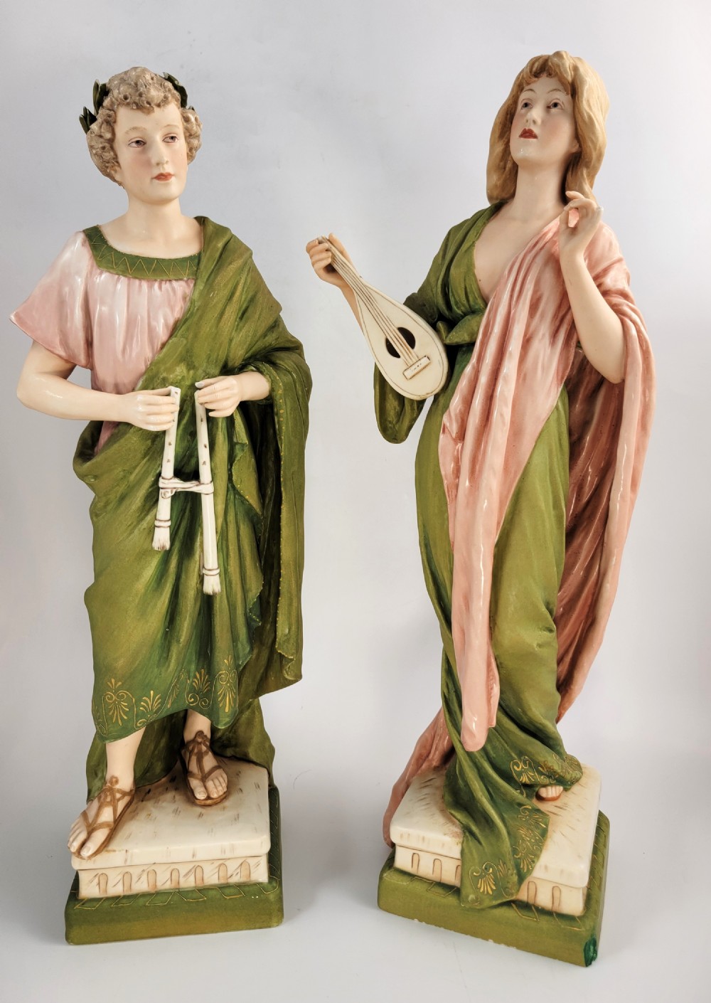 A pair of antique porcelain figures depicting a male and female in draped green and pink classical robes holding musical instruments.