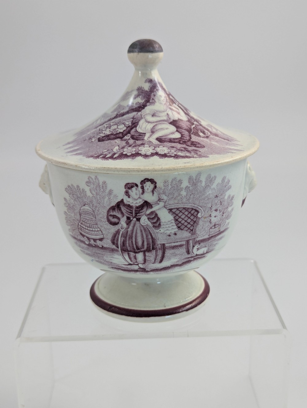 A 19th-century Staffordshire covered earthenware sugar bowl featuring a purple mulberry transferware romantic scene with lion mask handles and a pointed lid.