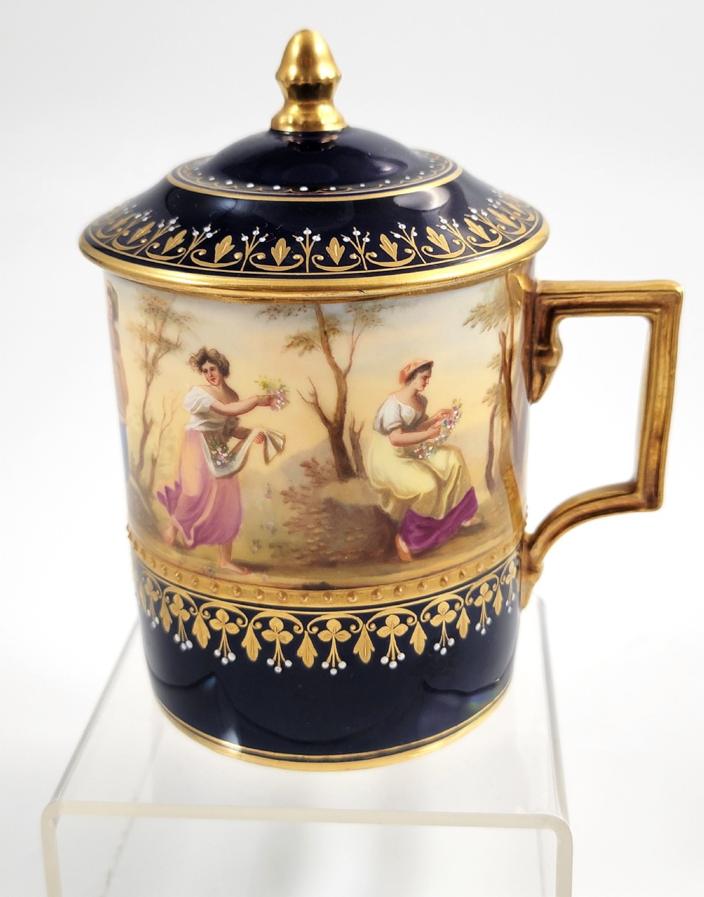 A cobalt blue porcelain covered cup featuring gold leaf scrollwork trim and a hand-painted allegorical woodland vignette with classical figures.