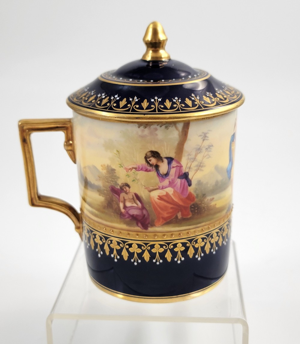 A cobalt blue porcelain covered cup featuring gold leaf scrollwork trim and a hand-painted allegorical woodland vignette with classical figures. View V