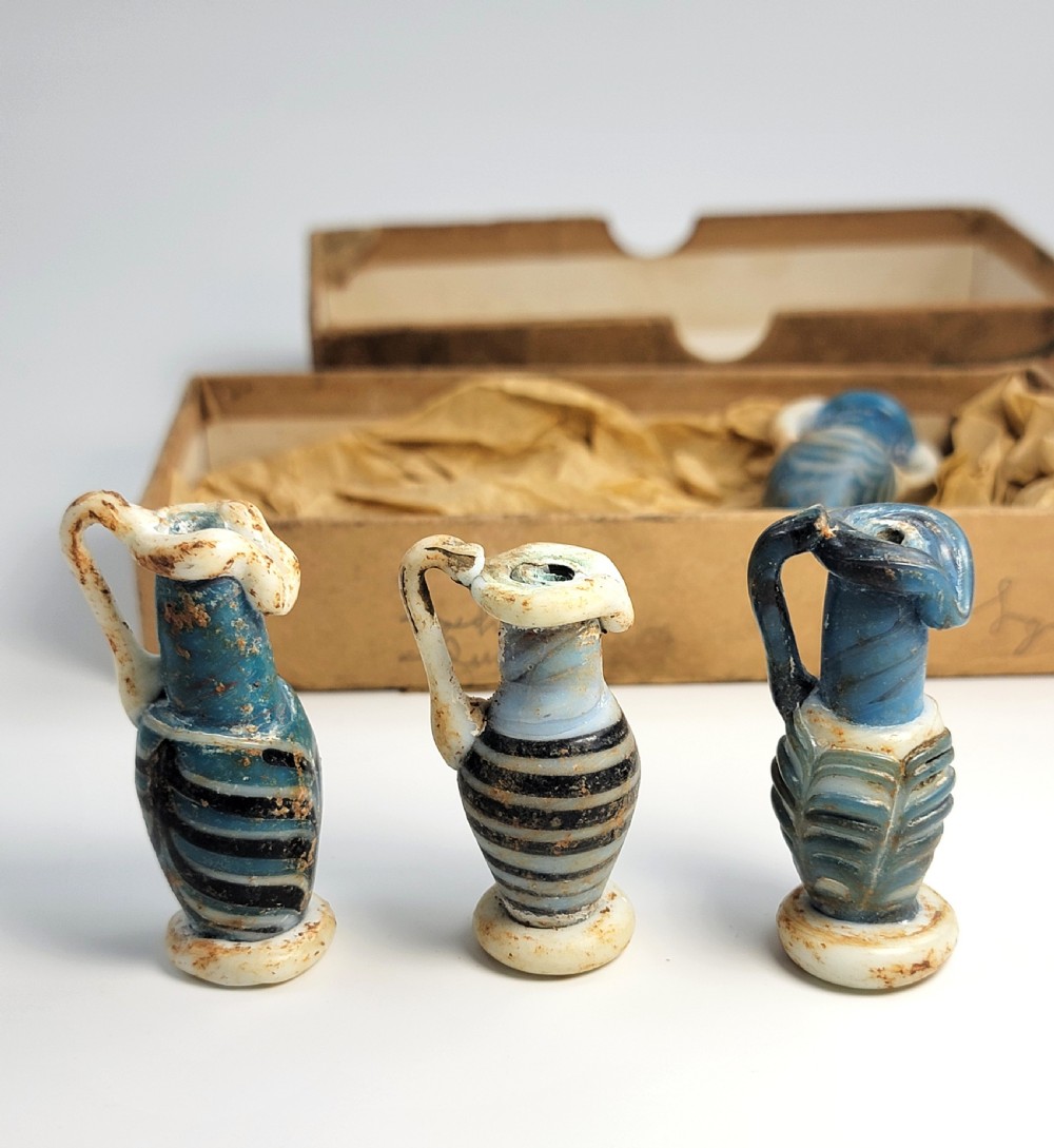 A set of four small ancient Roman glass unguentaria with blue bodies, white trailed decoration, and small handles resting inside a cardboard box. View III