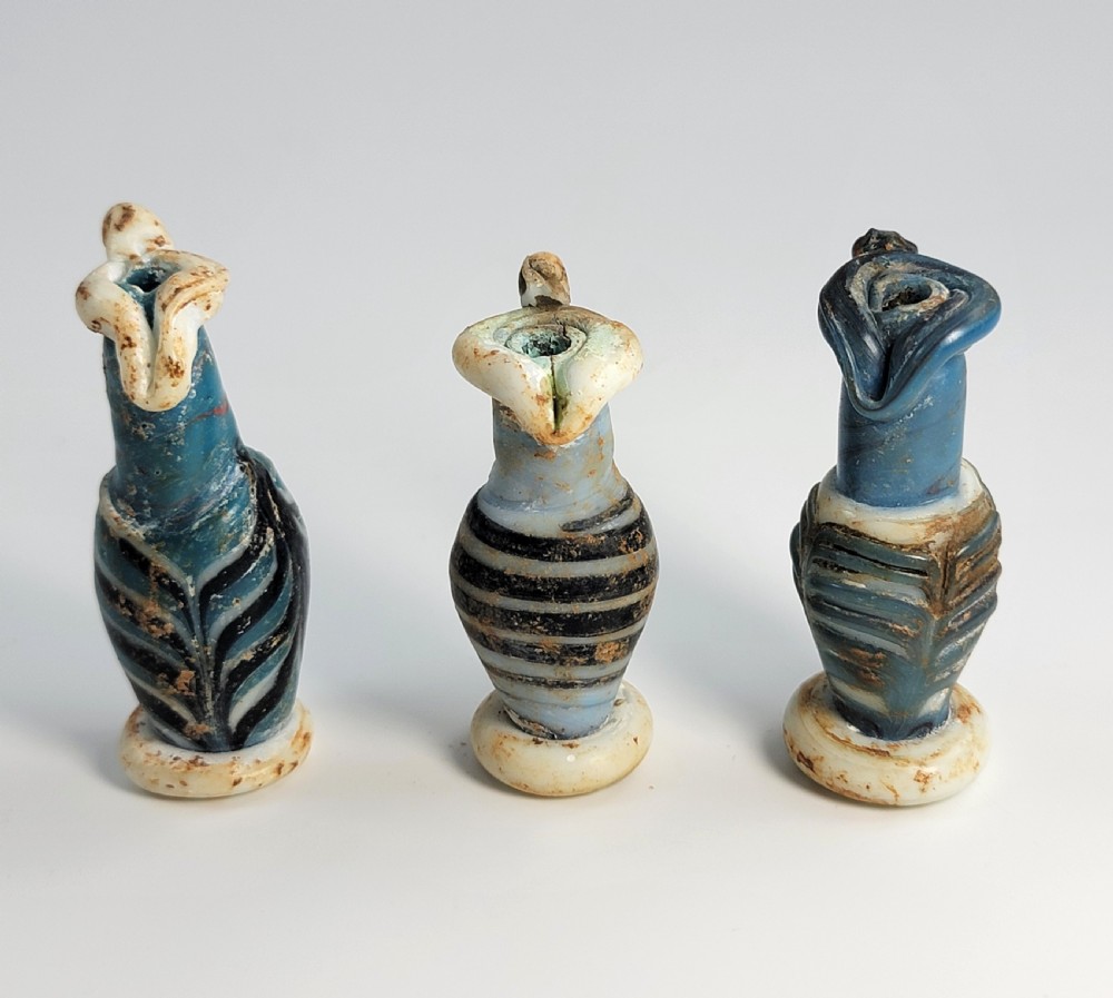 A set of four small ancient Roman glass unguentaria with blue bodies, white trailed decoration, and small handles resting inside a cardboard box. View IV